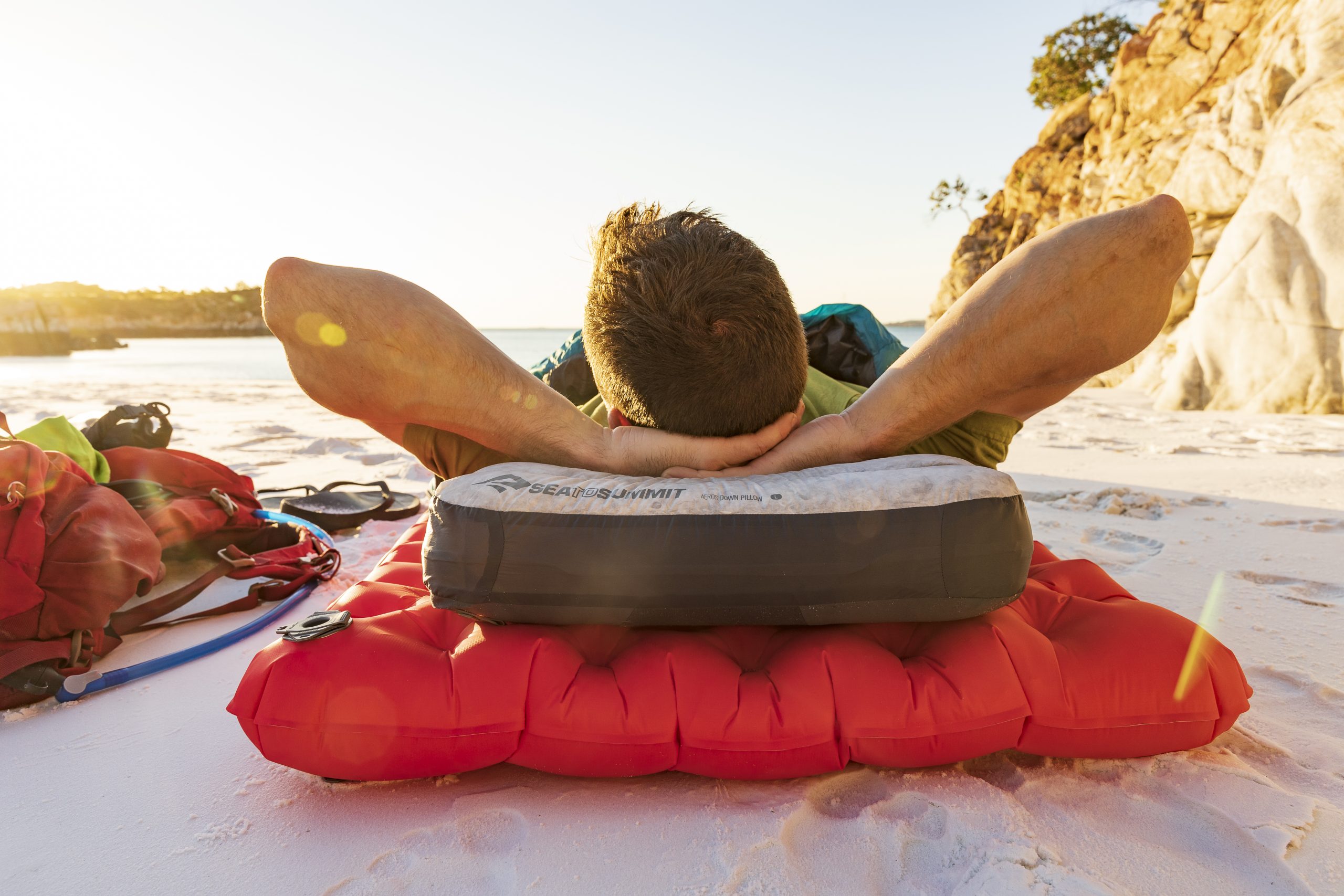 Sea to Summit offers all sorts of camp gear, including inflatable pillows and sleeping pads like the red pad and gray and white pillow shown here with a man laying on it.